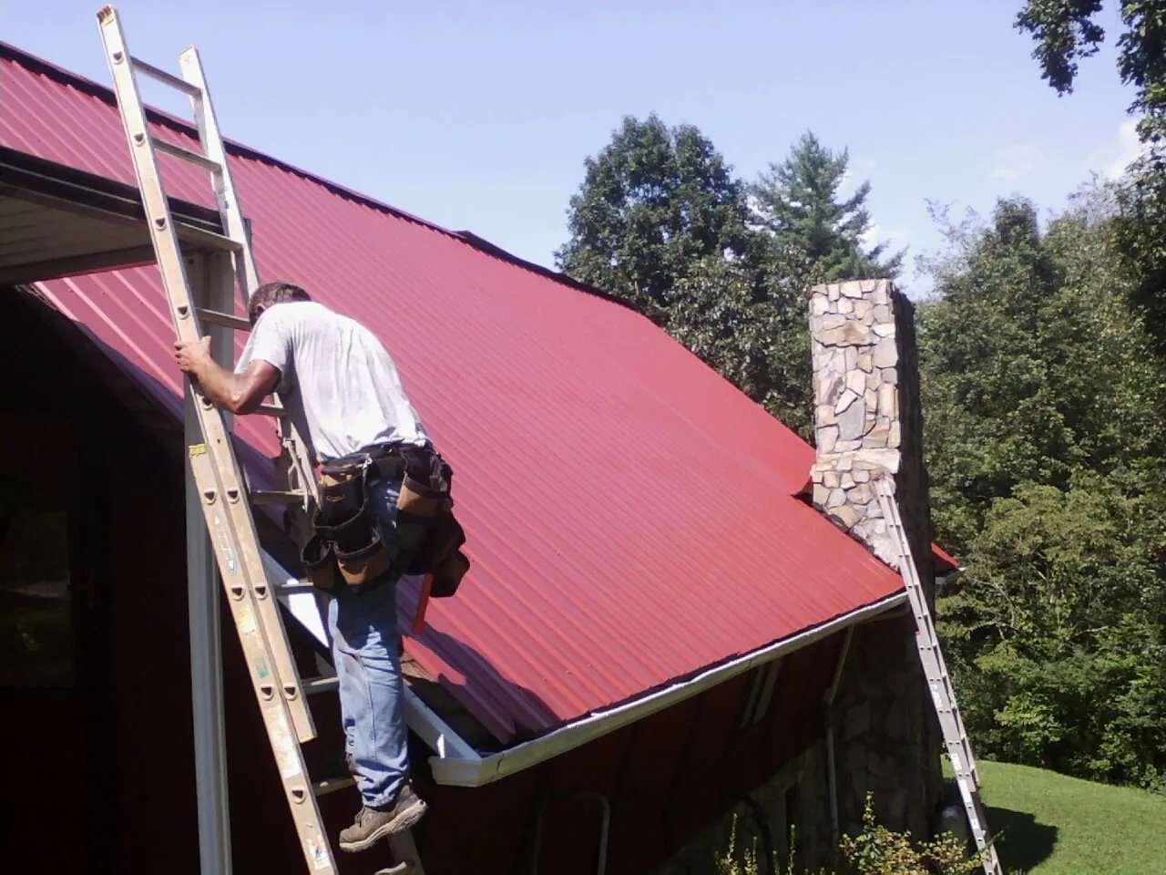 Roofer on a ladder beside a red metal roof and stone chimney — Soffit & Fascia Repair by Roofing Brothers in Russells Point
